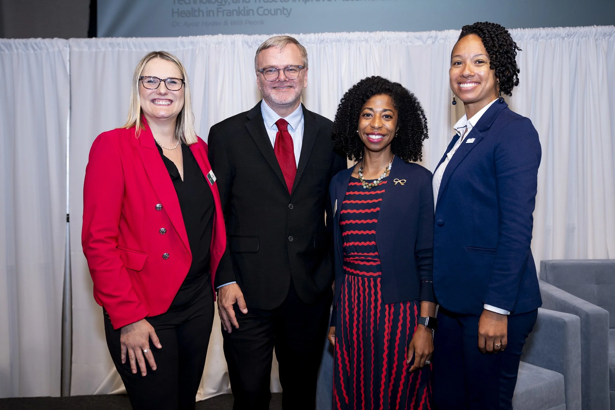 Group photo of four professionally dressed people smiling at a conference or event with white curtains and a presentation screen in the background.