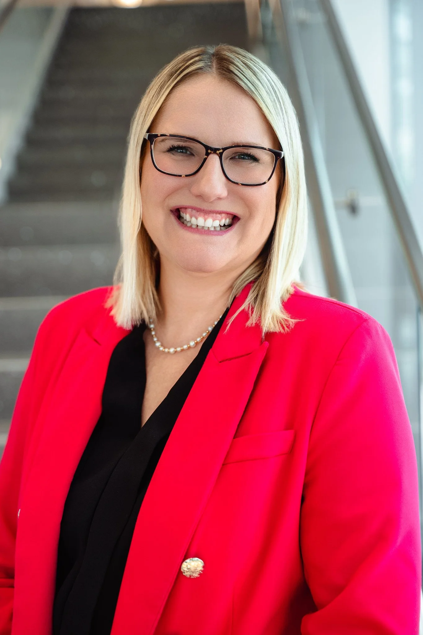 A smiling woman with blonde hair and glasses, wearing a red blazer and a pearls, standing in a modern building with stairs in the background.