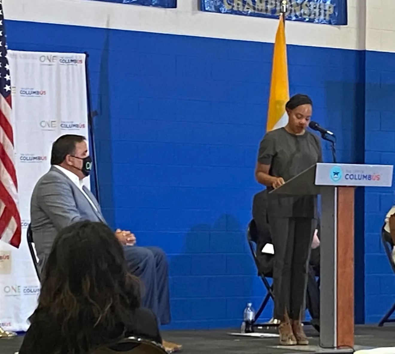 A woman speaking at a podium during a public event, with a blue wall and flags in the background, including an American flag and a yellow flag. A man seated nearby is wearing a gray suit and face mask.
