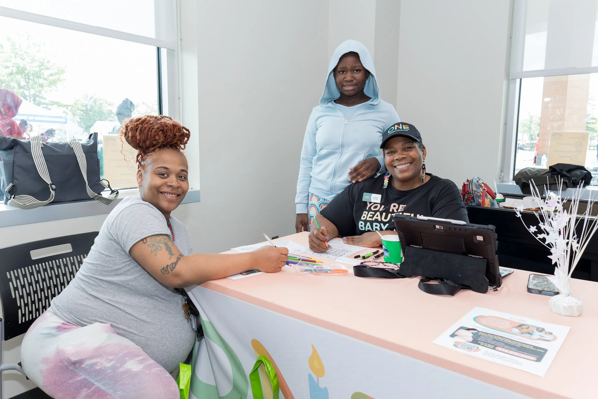 Three women sitting at a table inside a room with large windows, smiling at the camera. One woman is on the left with a gray shirt and colorful pants, the second woman is in the middle with a black shirt and a cap, and the third woman is standing behind them wearing a light blue hoodie.