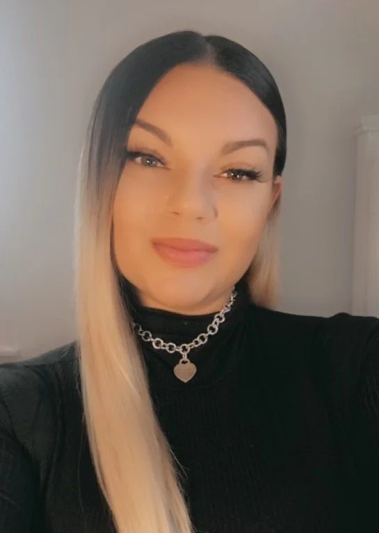 A woman with long blonde hair, wearing a black turtleneck and a silver chain necklace with a heart pendant.