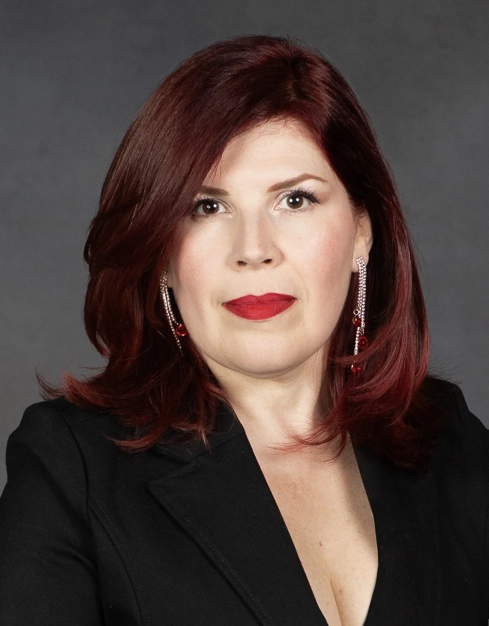 A portrait of a woman with red hair, red lipstick, and wearing earrings and a black blazer against a gray background.