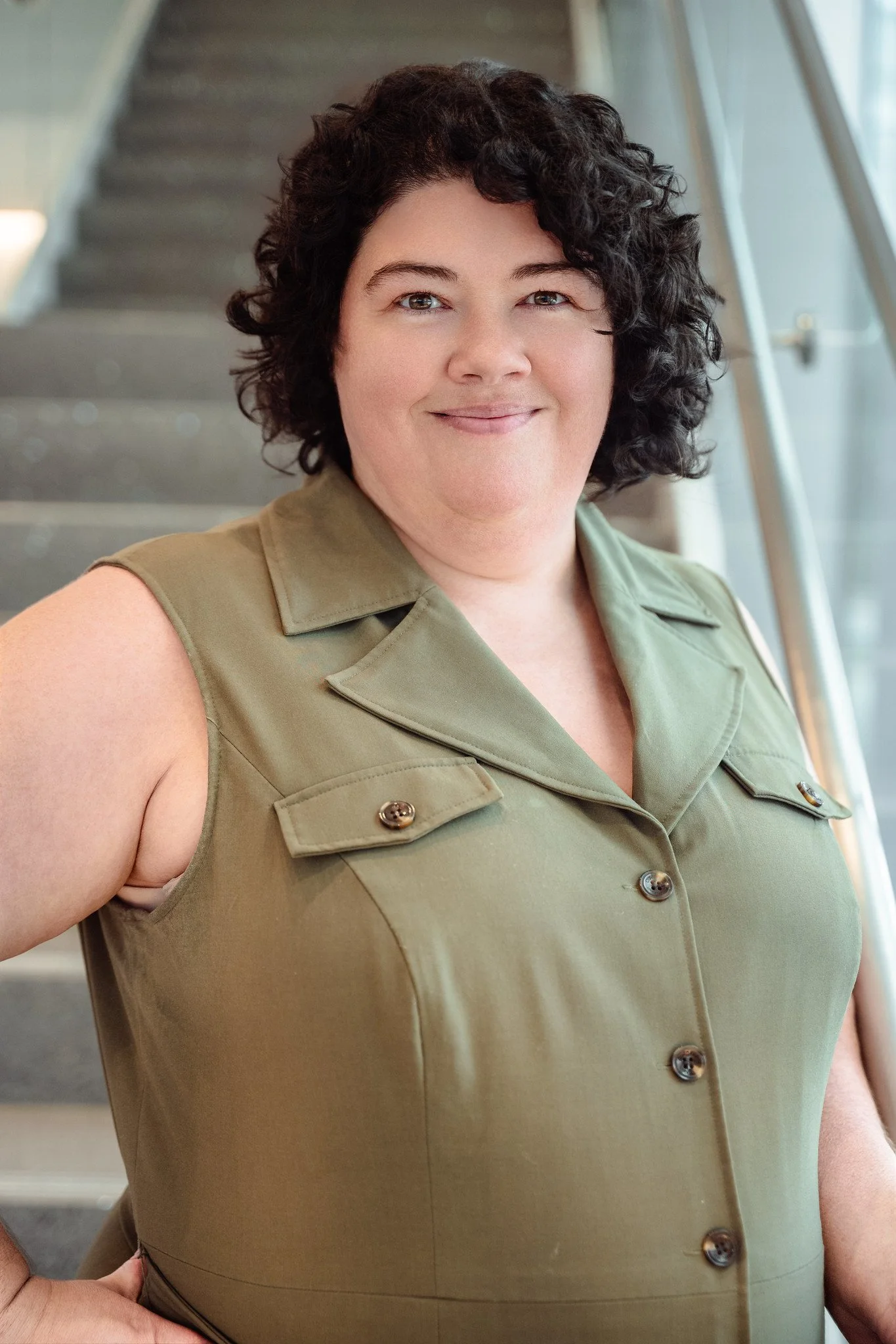 A woman with curly dark hair smiling, wearing an olive green sleeveless button-up shirt, standing indoors near a staircase and glass railing.