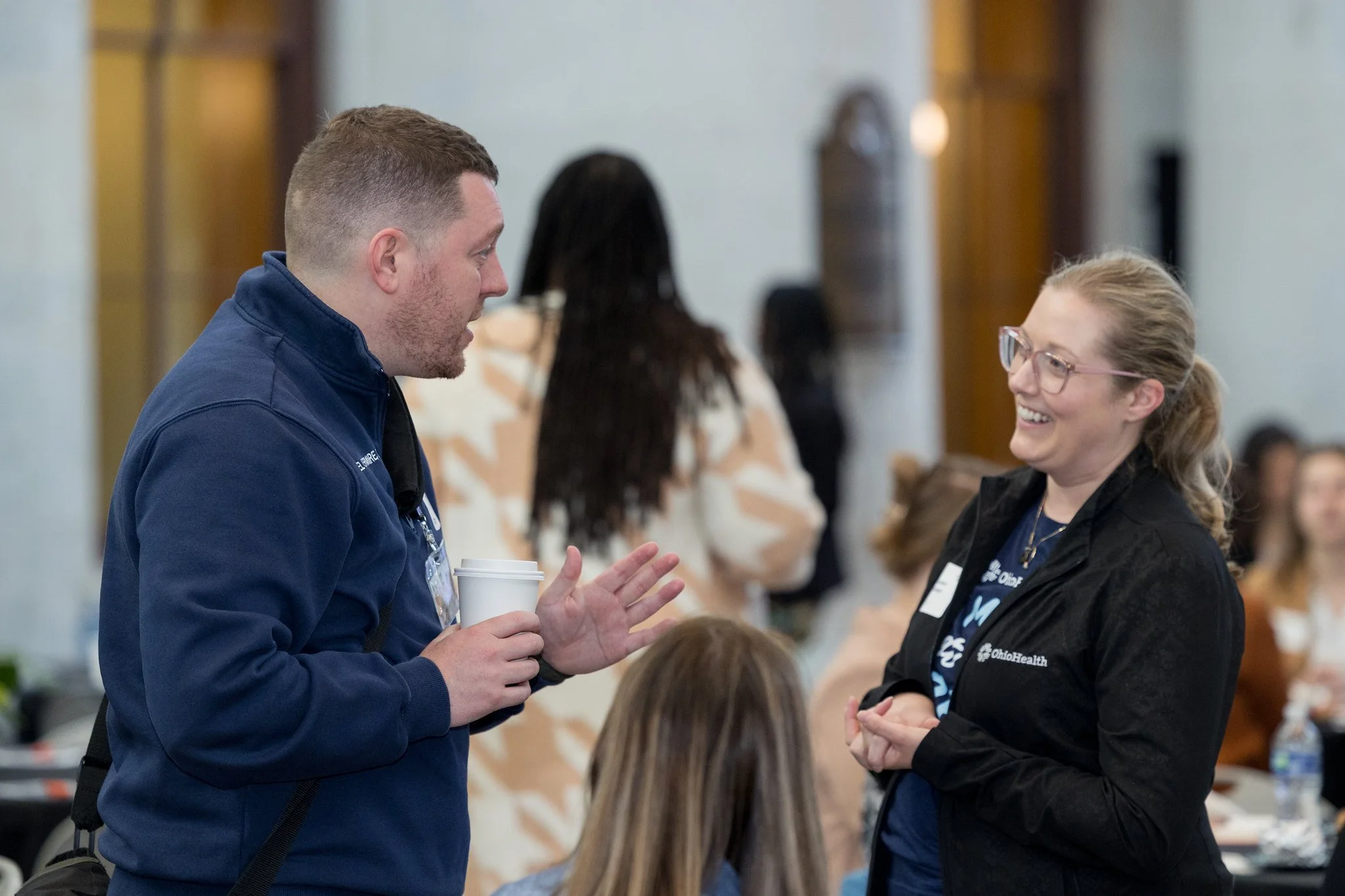A man and a woman are engaged in conversation indoors. The man is wearing a blue jacket and holding a disposable coffee cup, while the woman has glasses and is smiling, dressed in a black jacket with an OhioHealth logo. They are surrounded by other people in a busy setting, possibly a conference or social event.