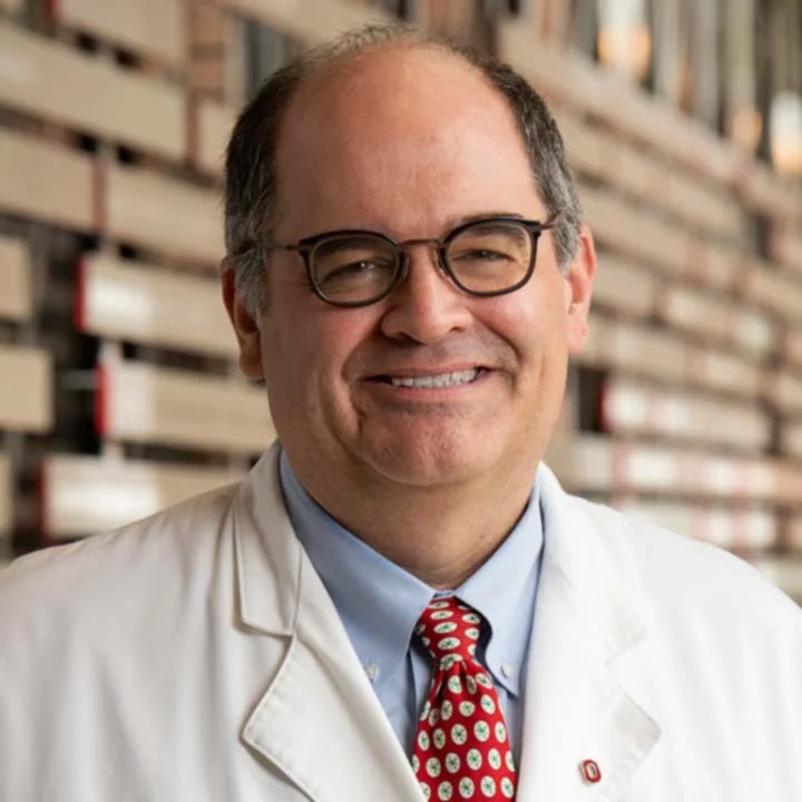A middle-aged man with glasses, smiling, in a white lab coat, standing in front of a wooden wall. He is wearing a blue shirt and a red patterned tie.