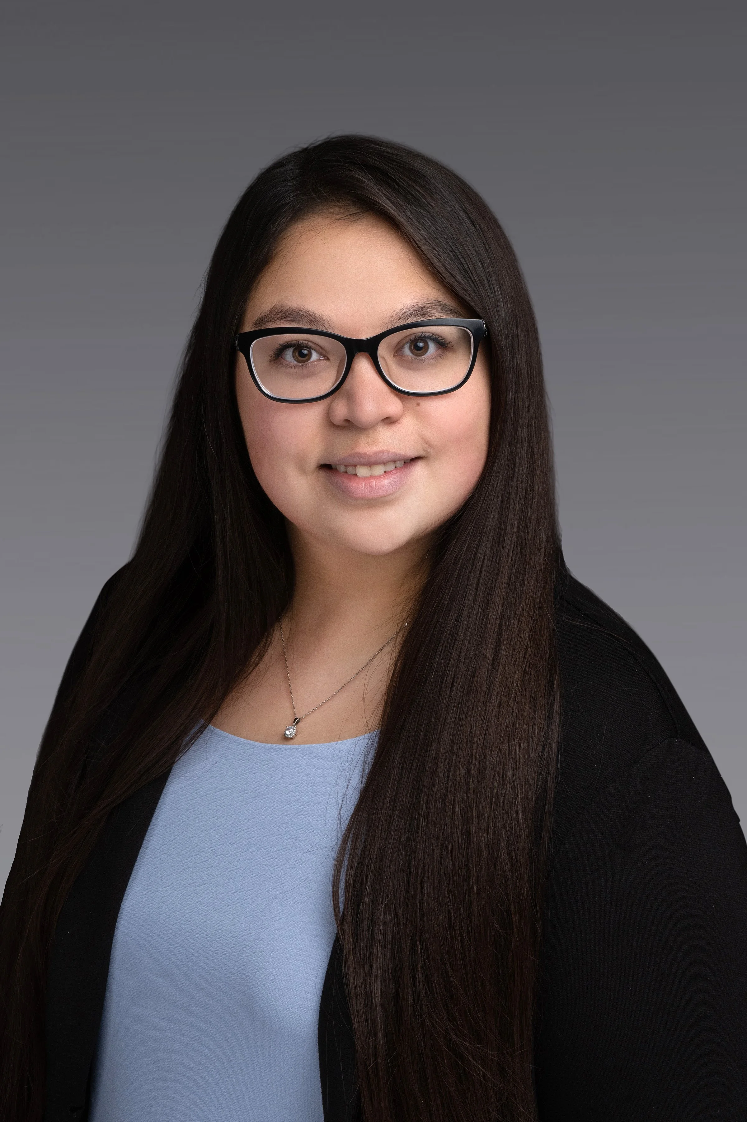 Professional headshot of a woman with long dark hair, glasses, a black blazer, light blue top, and a silver necklace against a gray background.