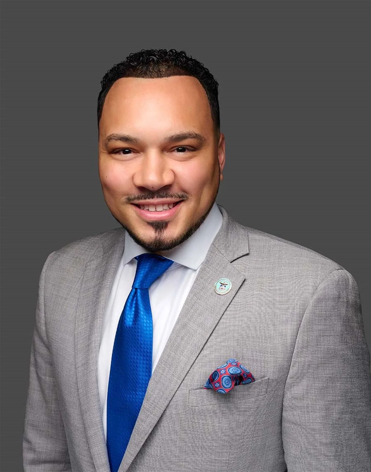 A man in a gray suit with a blue tie and a colorful pocket square, smiling against a solid gray background.