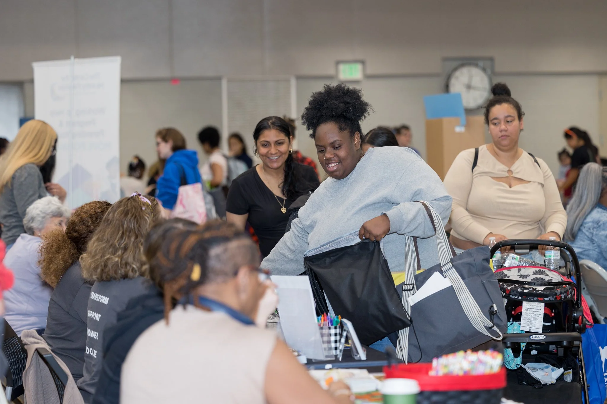 A group of people at a community event or gathering, with some engaging at tables and others standing, in an indoor setting with a clock on the wall.