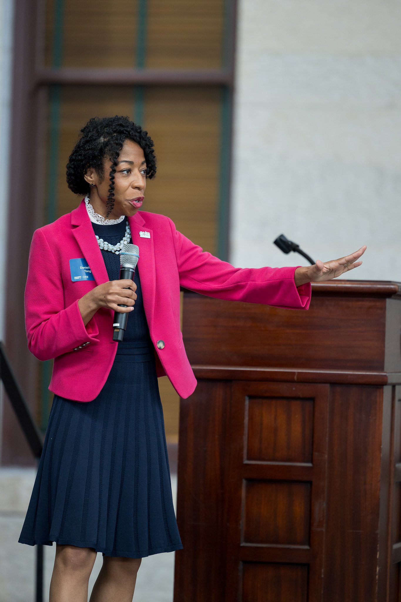 A woman wearing a pink blazer and navy dress giving a speech at a podium, holding a microphone in her right hand and gesturing with her left hand.