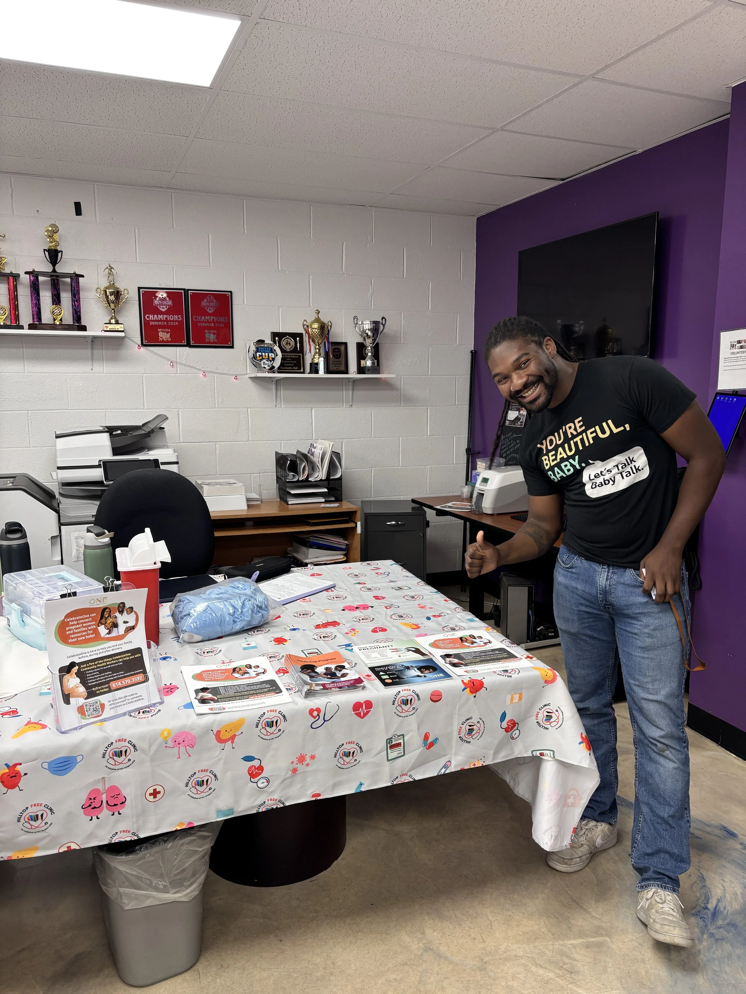 Man smiling and giving a thumbs up in a room with a table covered with health-related flyers and supplies. Shelves with trophies and framed awards are on the wall behind him.