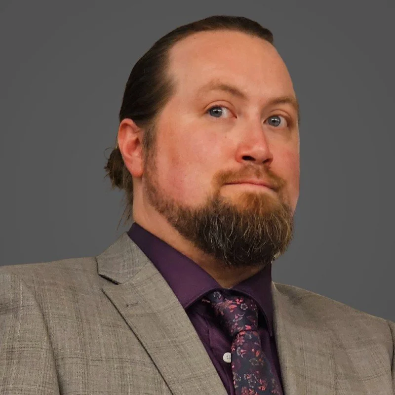 A man with a full beard in a suit with a blazer and a purple shirt, wearing a tie, looking confident against a plain gray background.
