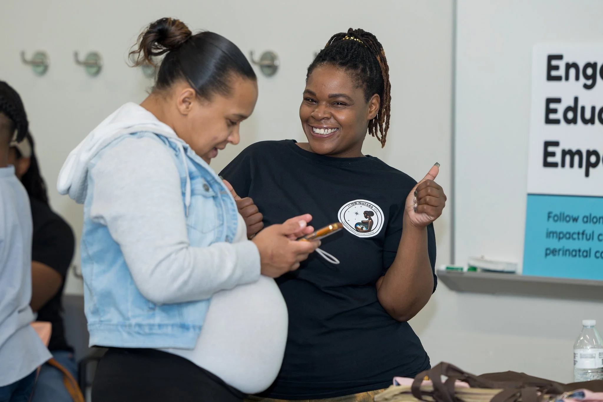 Two women, one pregnant, engaging in a conversation, smiling, in a classroom or seminar setting with a poster in the background.