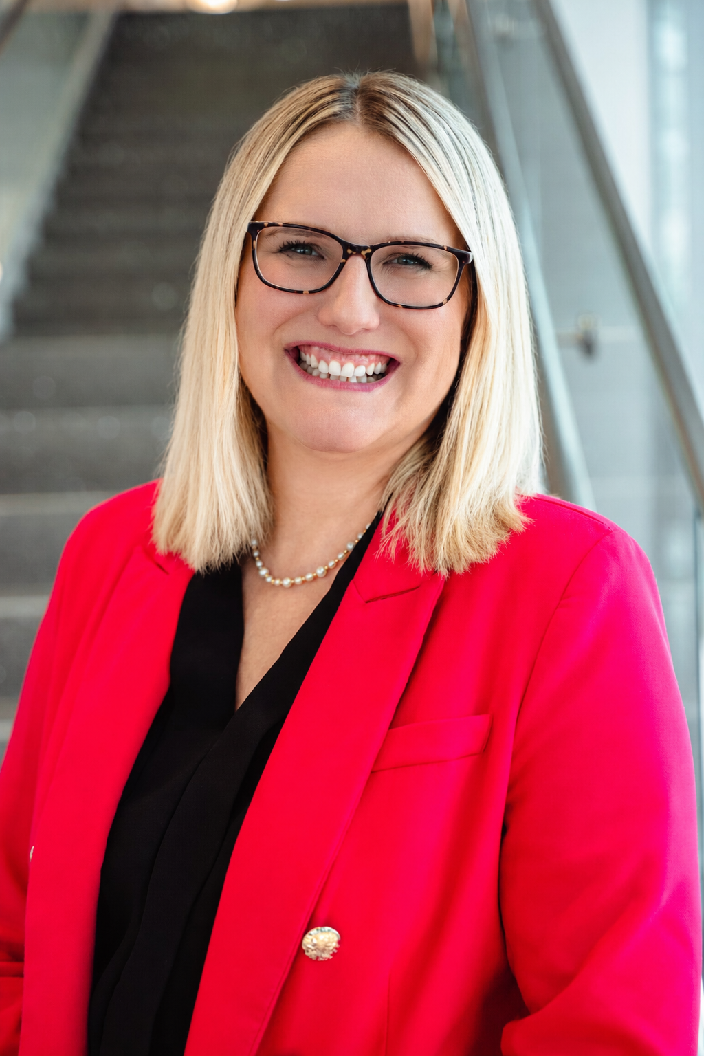 A smiling woman with blonde hair and glasses, wearing a red blazer and a pearls, standing in a modern building with stairs in the background.