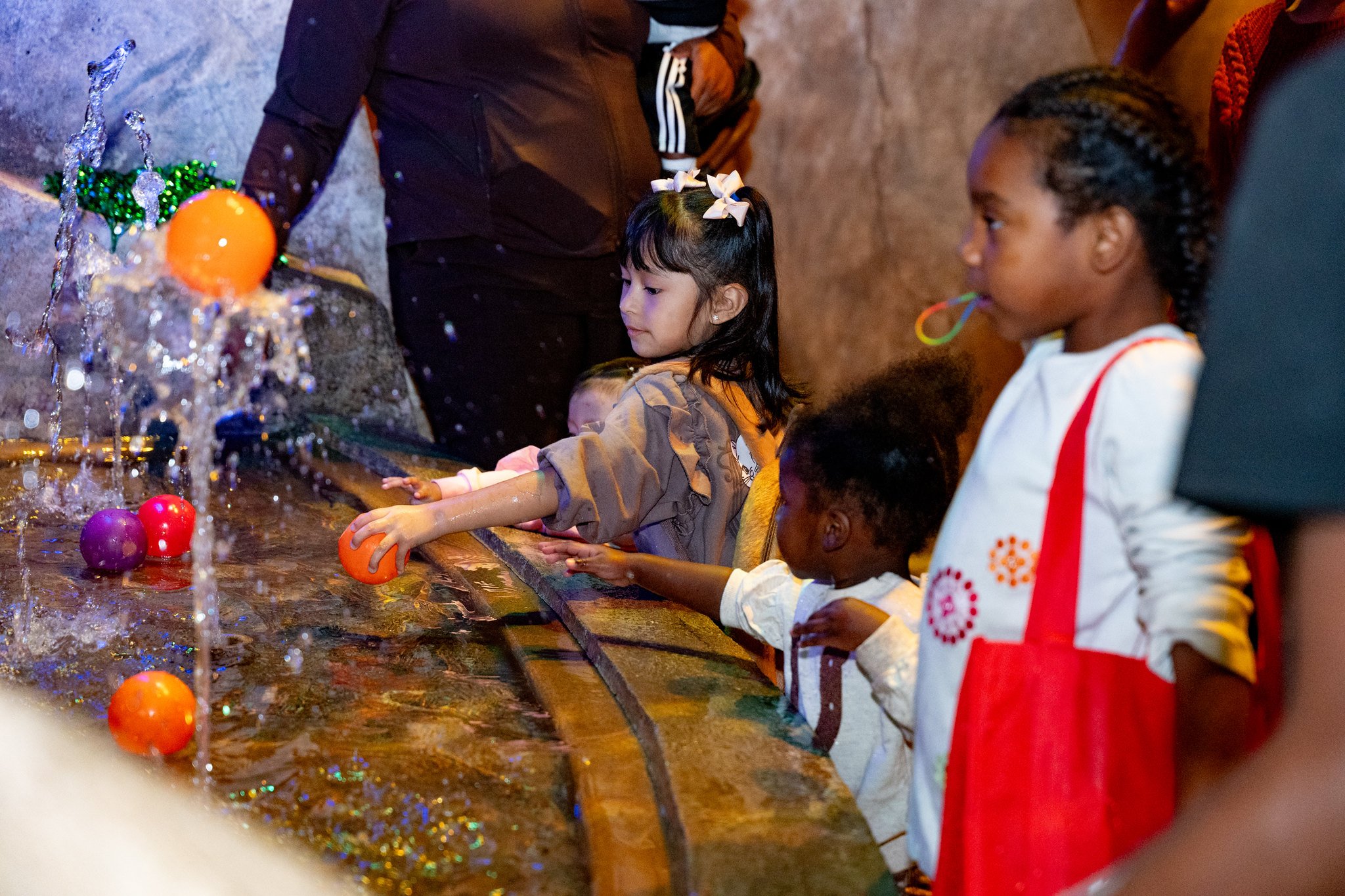 Children and adults playing a water ring toss game with floating colorful balls at an indoor location with brown walls.