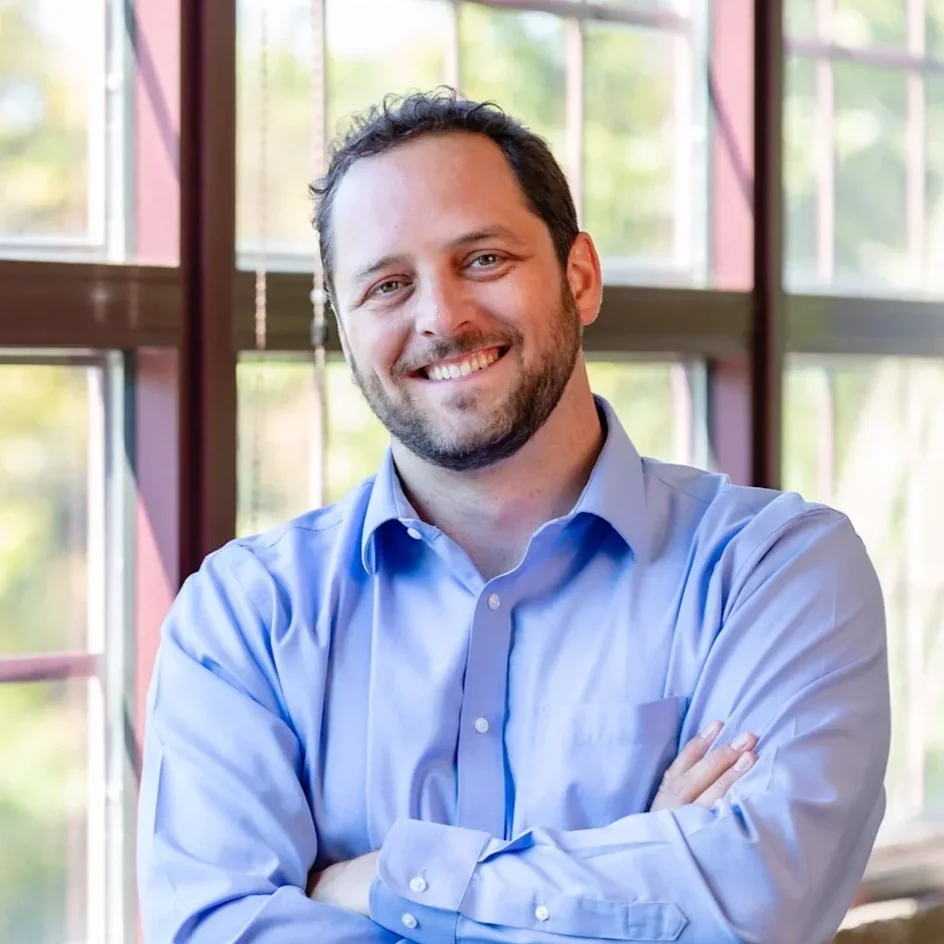 A man smiling with crossed arms, standing in front of large windows with a view of trees outside.