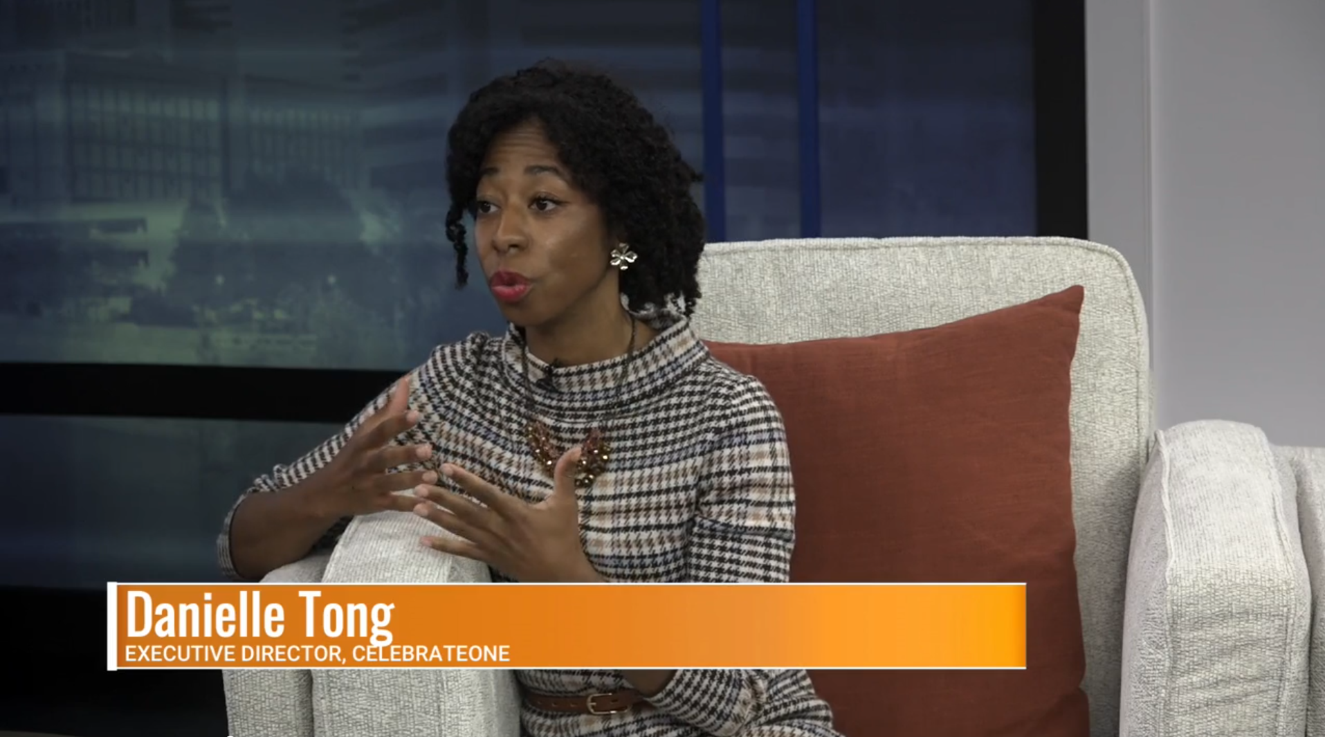 A woman with dark curly hair and red lipstick is speaking or explaining, seated on a light-colored armchair with a rust-colored pillow, wearing a checkered top and jewelry, with a cityscape background visible through a window.