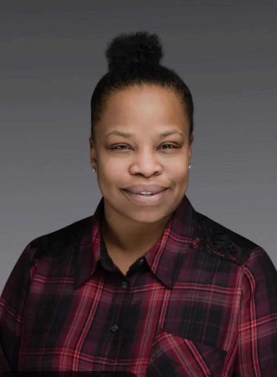 A woman with dark hair in a high bun, smiling slightly, wearing a red and black plaid shirt, against a gray background.