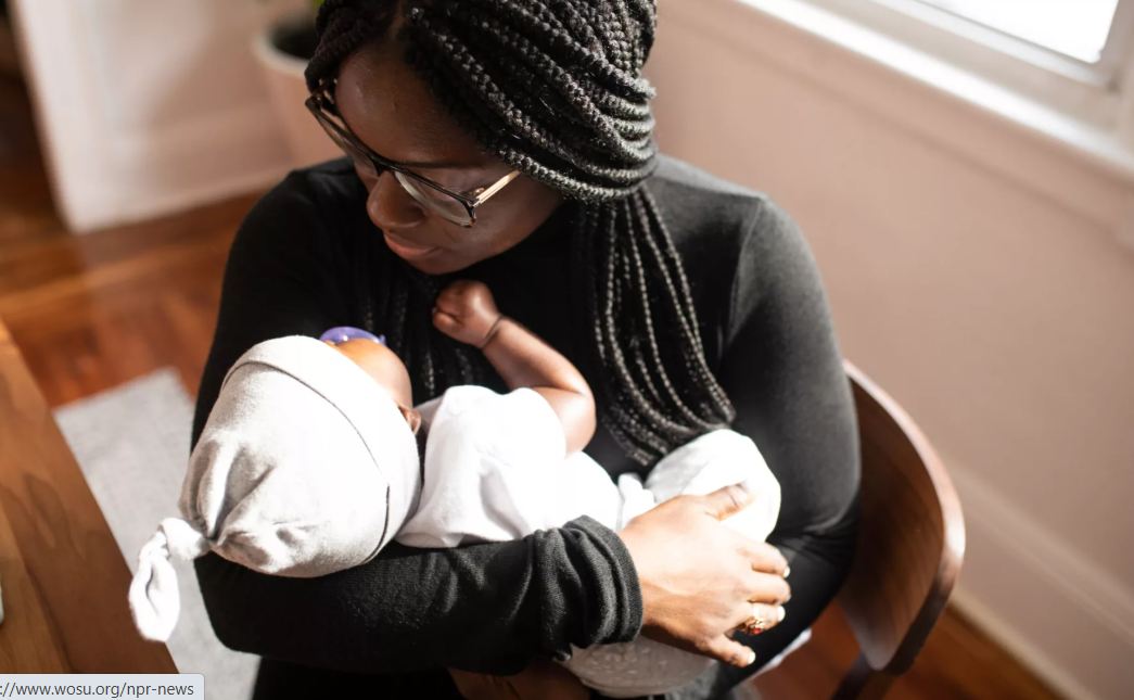 A woman with braided hair and glasses cradles a baby in her arms, looking down at the infant in an indoor setting with wooden floors and a window in the background.