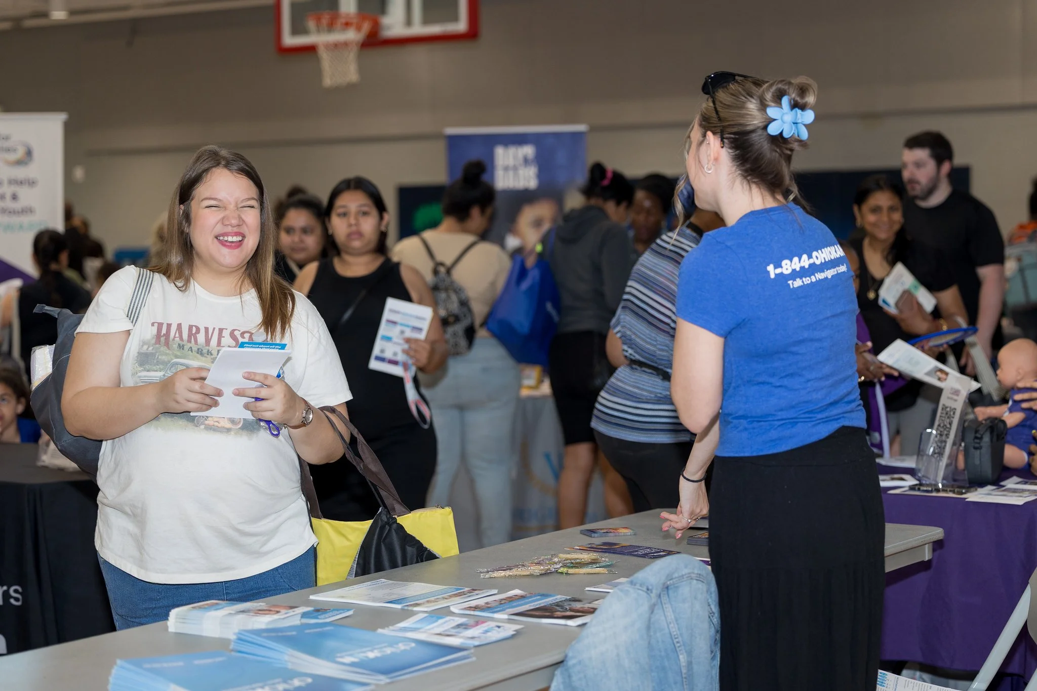 A woman smiling and showing her teeth at a booth during a health fair or conference, with a group of people in the background.