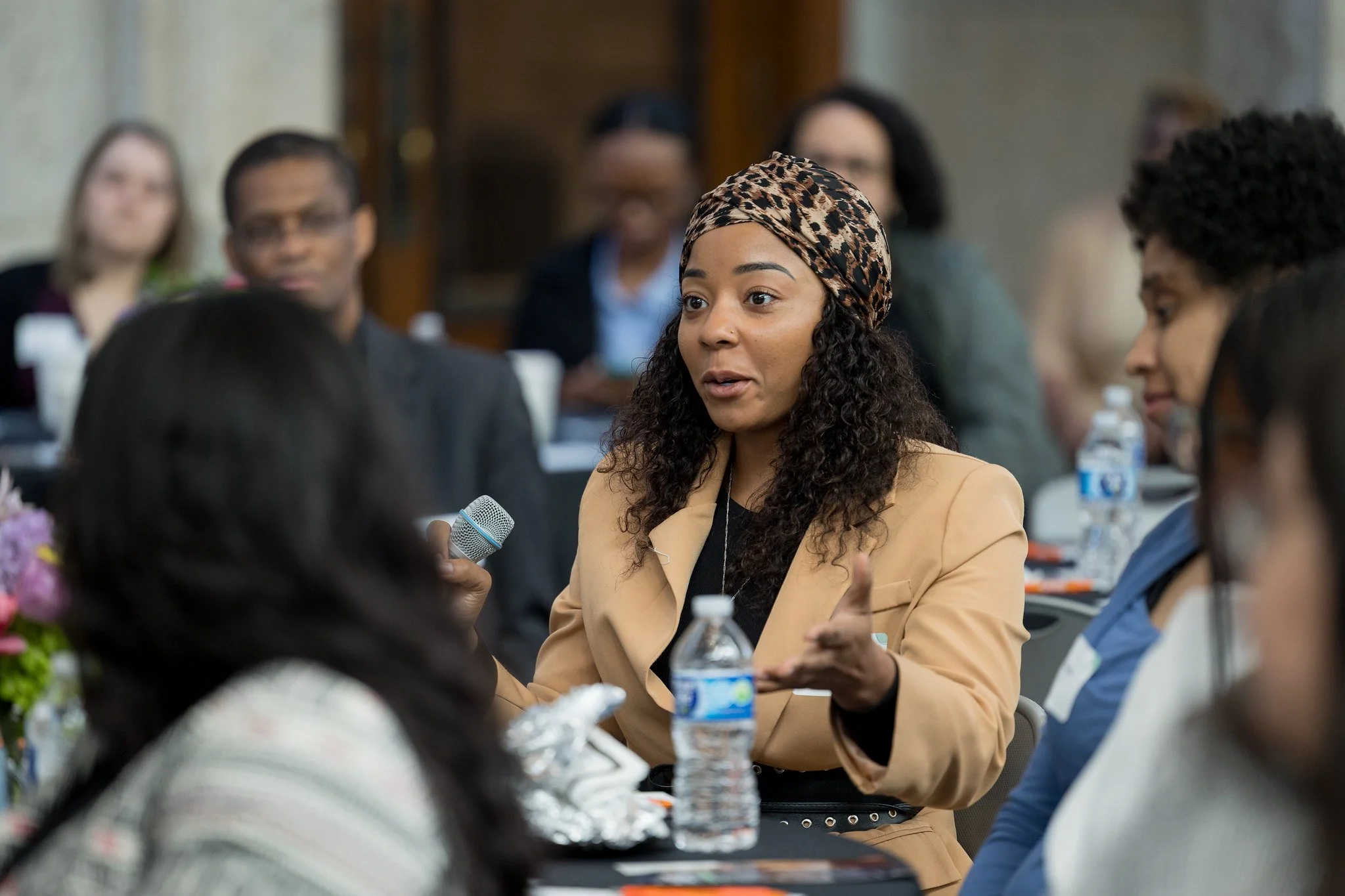 A woman with curly hair and a leopard print headscarf speaking into a microphone in a conference room, surrounded by other attendees.