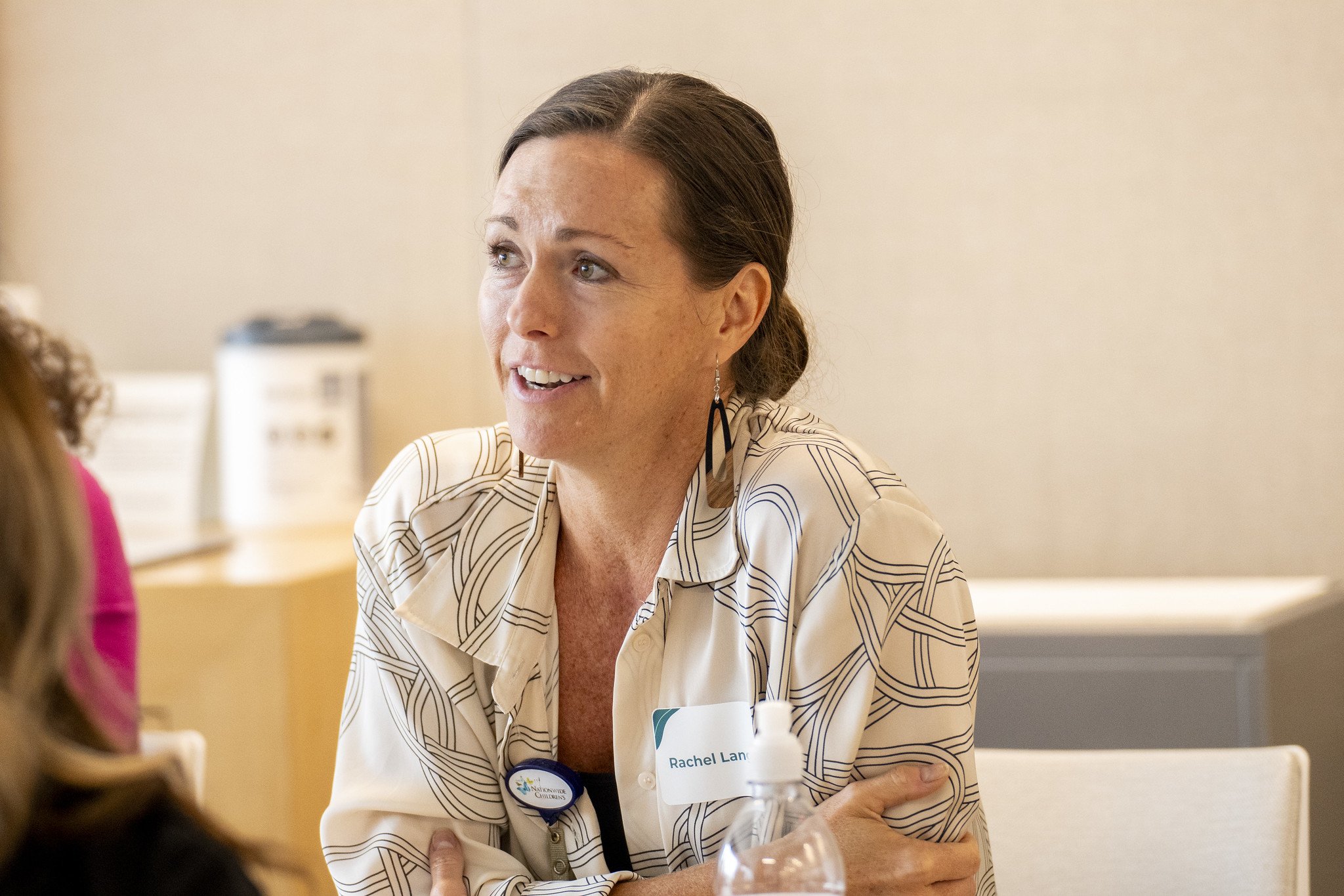 A woman with brown hair in a bun, wearing a patterned beige shirt and earrings, speaking with someone at a meeting or event.