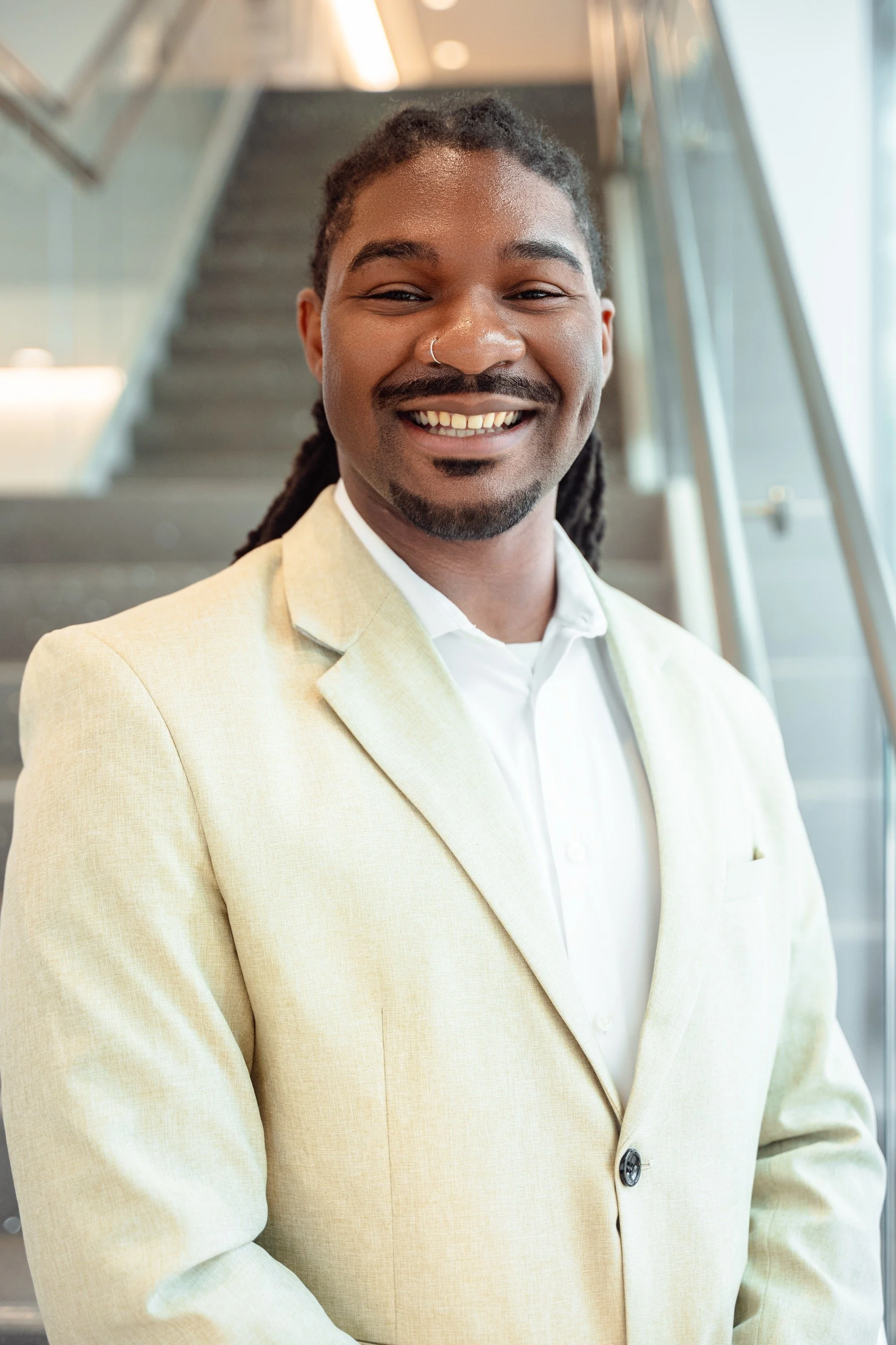 Portrait of a smiling man with dreadlocks, wearing a beige blazer and white shirt, standing inside a modern building with large glass windows and stairs in the background.