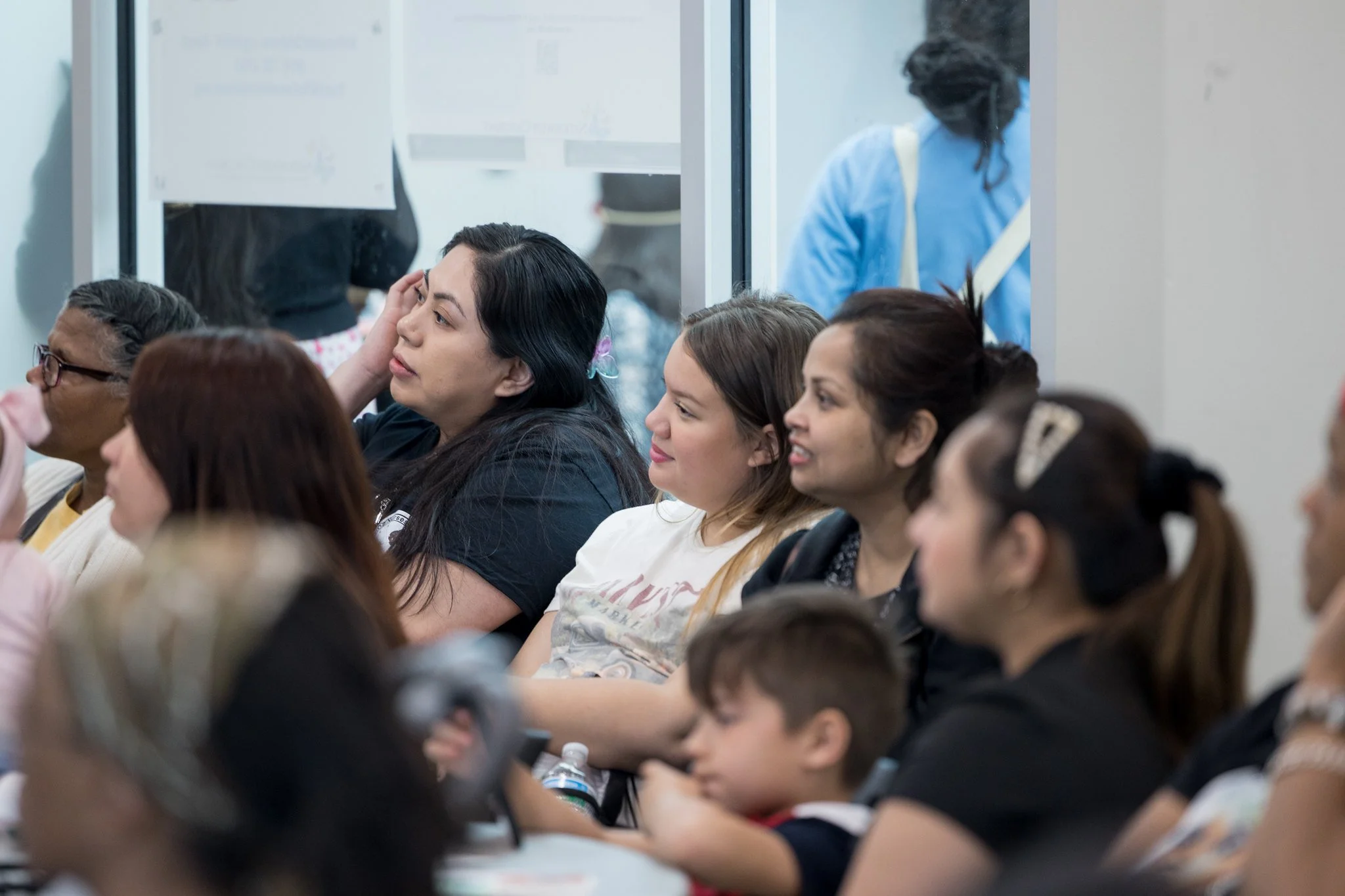 People seated in a waiting area, diverse in age and ethnicity, some with headphones, with a glass window behind them showing more people standing.