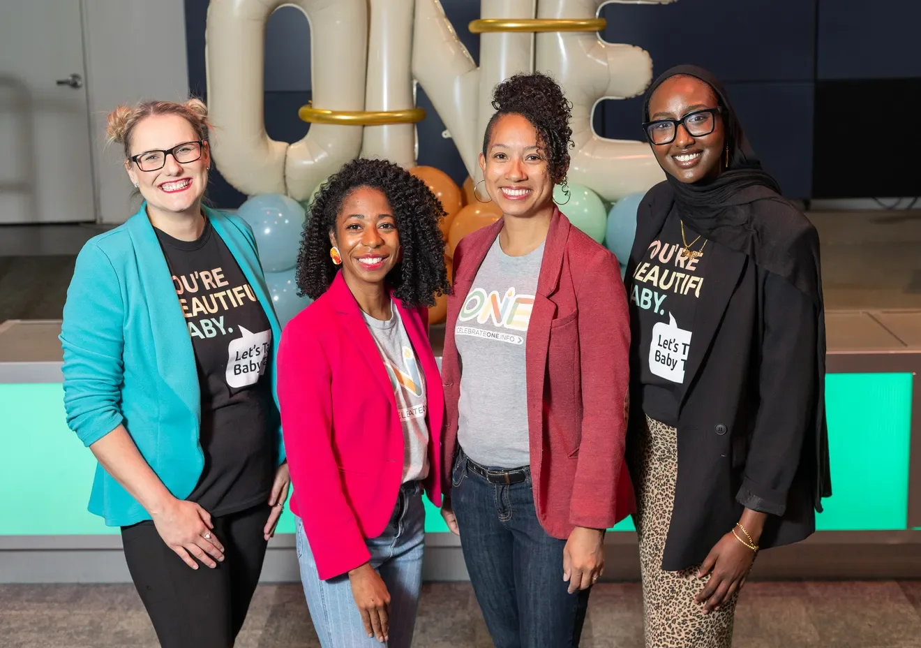 Four women standing together at an event, with balloons and ONE shaped decorations in the background, smiling at the camera.