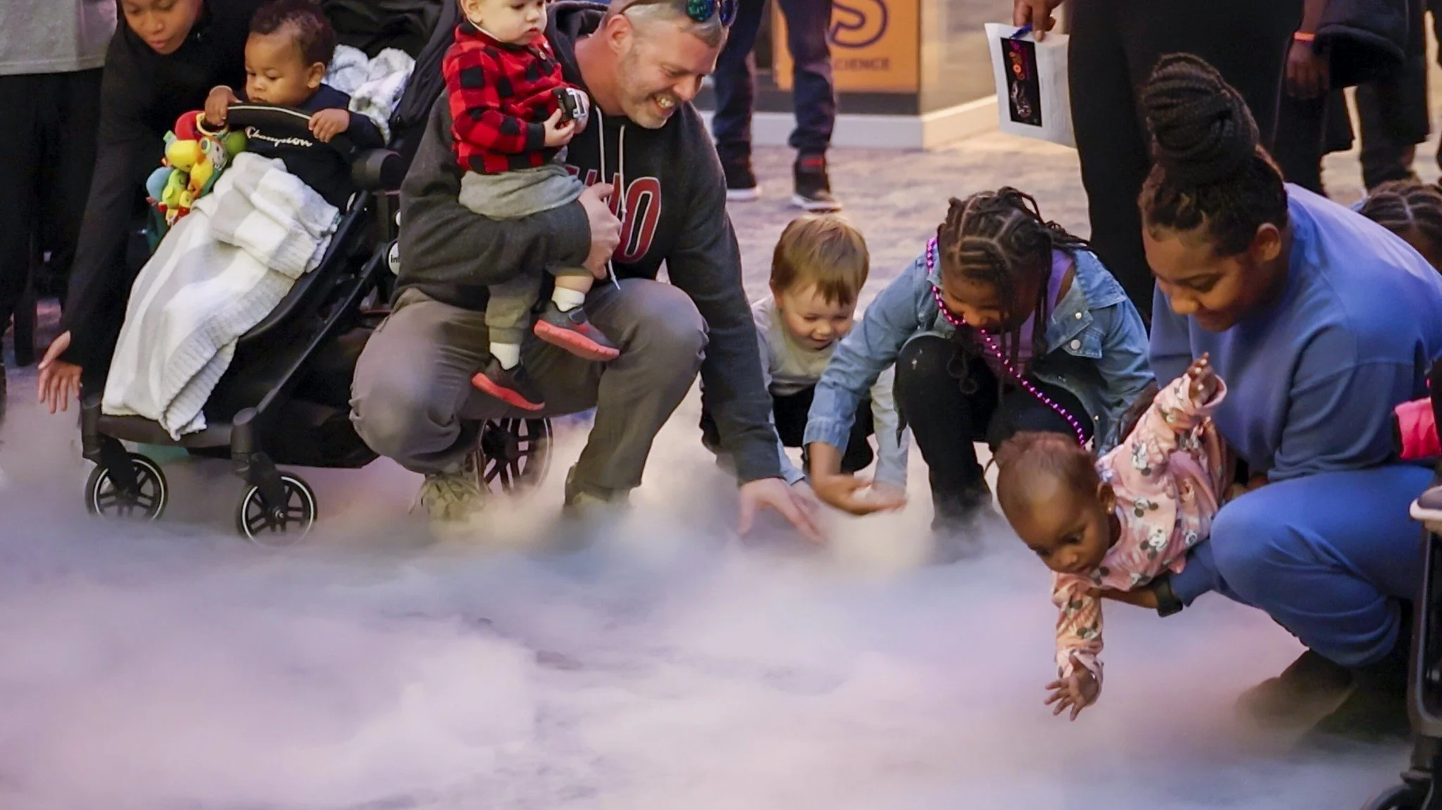 Group of children and adults gathered around and touching a fog or mist on the floor in an indoor setting, engaging with the mysterious material.