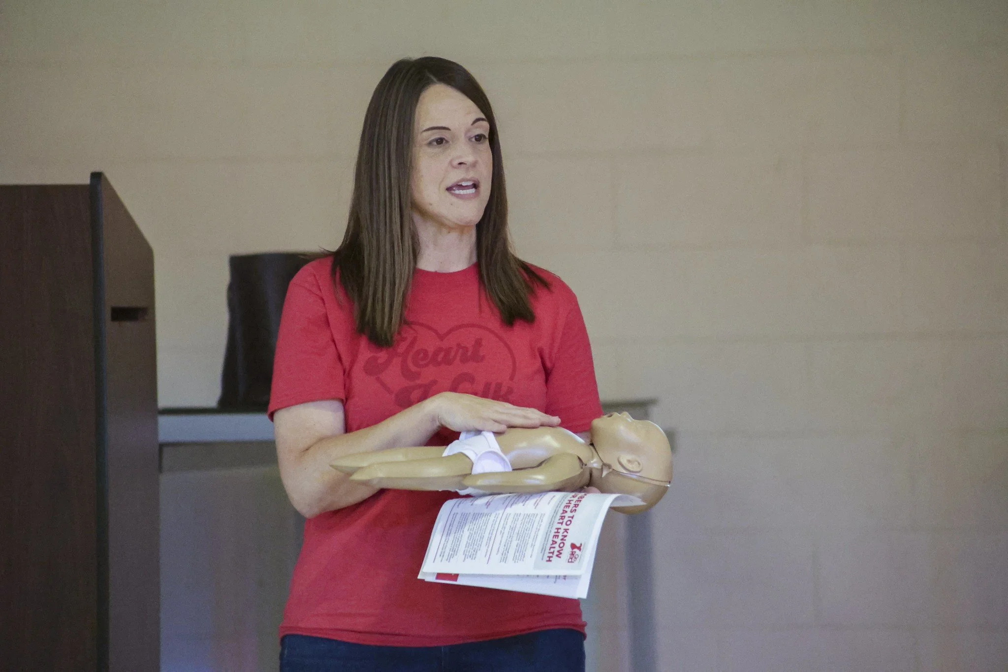 A woman in a red shirt holds a CPR Dummy Baby with her hand on the dummy's chest.