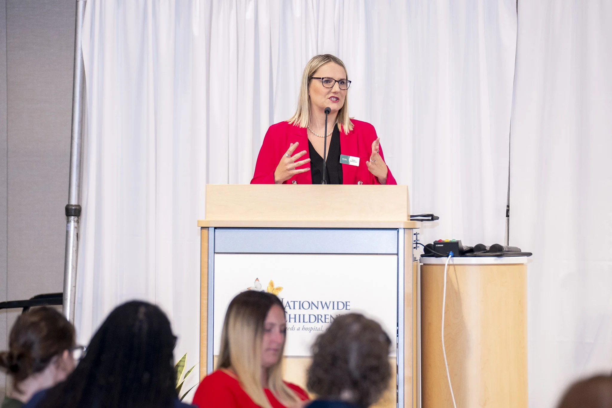 A woman in a red blazer giving a speech at a podium during a conference or event, with a mouth open and hands gesturing, in front of a white curtain and an audience.