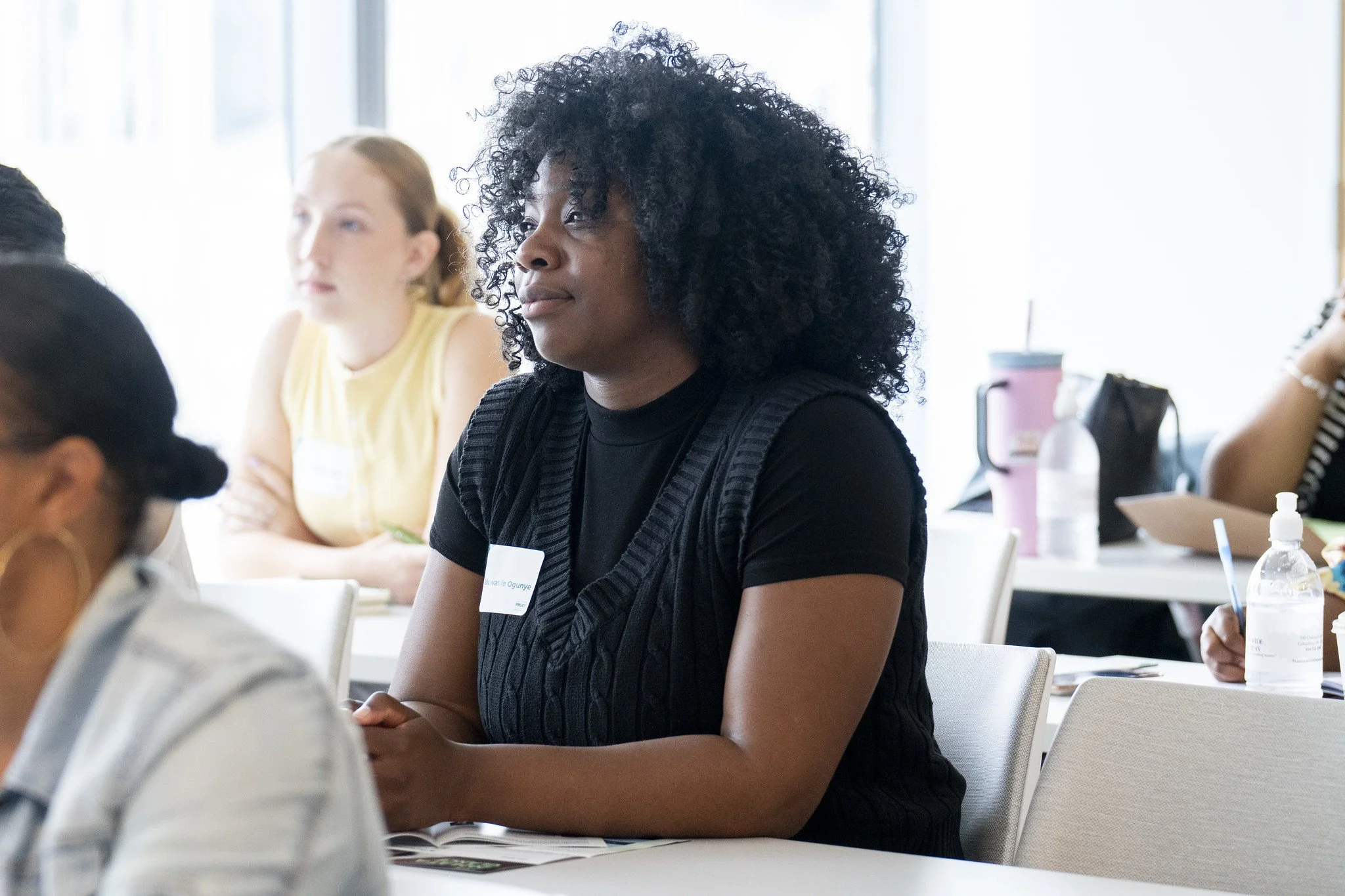 Woman with curly black hair, wearing a black shirt and a name tag, sitting at a table in a meeting or conference room with other women in the background.