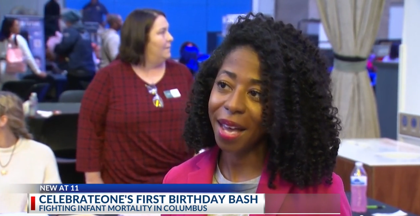A woman with curly hair wearing a pink blazer being interviewed at a celebration event for a child's first birthday with people in the background.