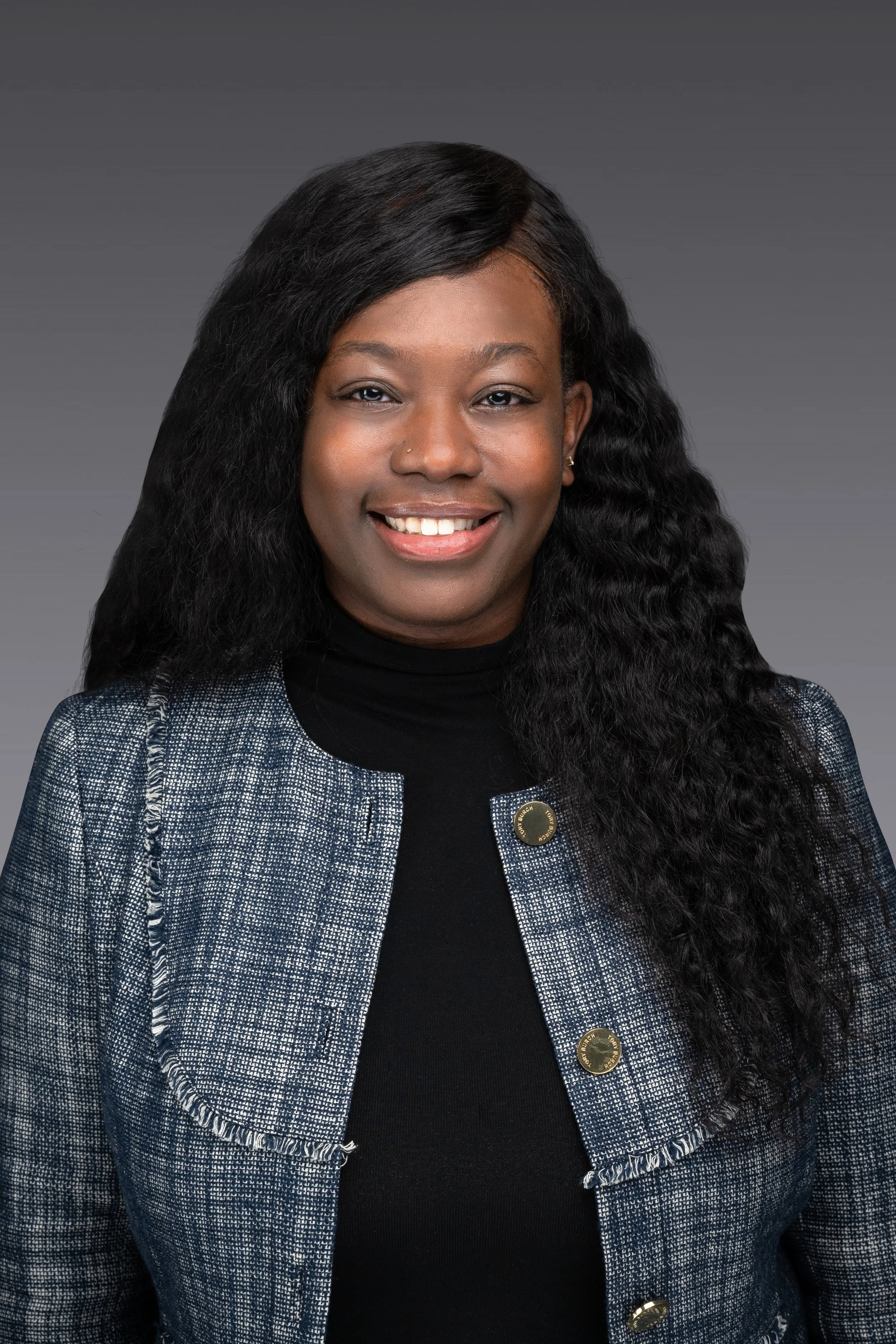 Professional portrait of a woman with long, wavy black hair, wearing a black top and a textured blue blazer, smiling at the camera against a gray background.