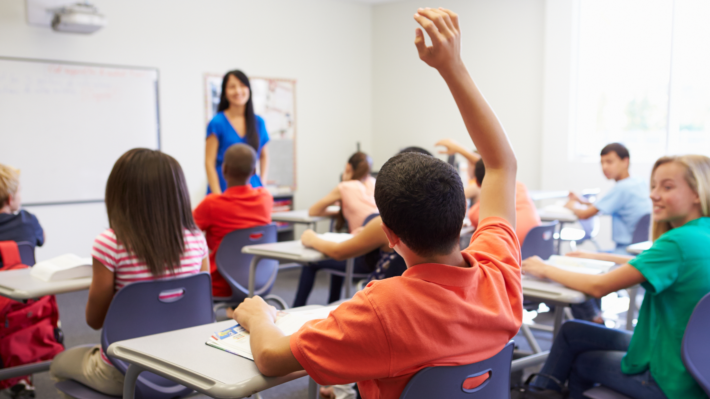 Students in a classroom with one student raising his hand, facing a teacher at the front of the room, with others sitting at desks.