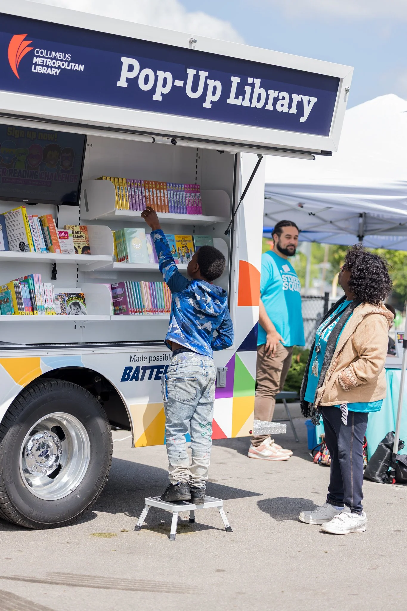 A boy standing on a small stool at a mobile library truck, reaching for books on a top shelf while two adults watch nearby. The truck has a sign that reads 'Pop-Up Library' and features colorful graphics and books displayed inside.