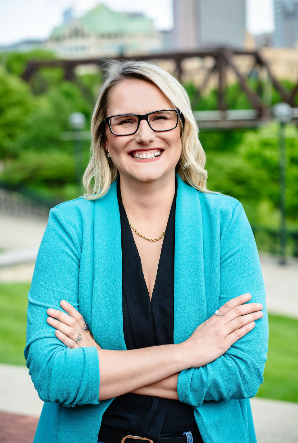 A smiling woman with blonde hair and glasses, wearing a red blazer and a pearls, standing in a modern building with stairs in the background.