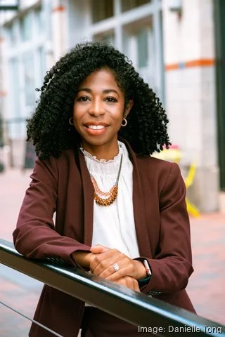A woman with curly black hair smiling, wearing a white blouse and a brown blazer, standing outdoors in an urban setting.