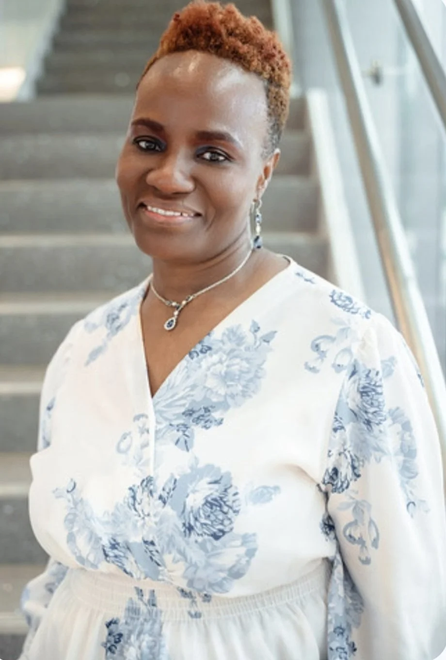 A woman with short brown hair, wearing a floral white and blue dress, jewelry, and smiling, standing on stairs near a glass railing.