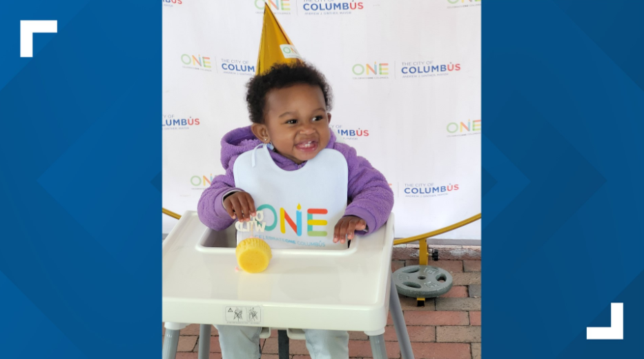 A smiling young girl sitting in a white high chair, wearing a purple hoodie and a white bib with the colorful text 'ONE.' She is holding a yellow toy or object with her right hand. Behind her is a backdrop with the words 'The City of Columbus' and 'ONE' logo, and a yellow cone-shaped party hat sticker above her head.