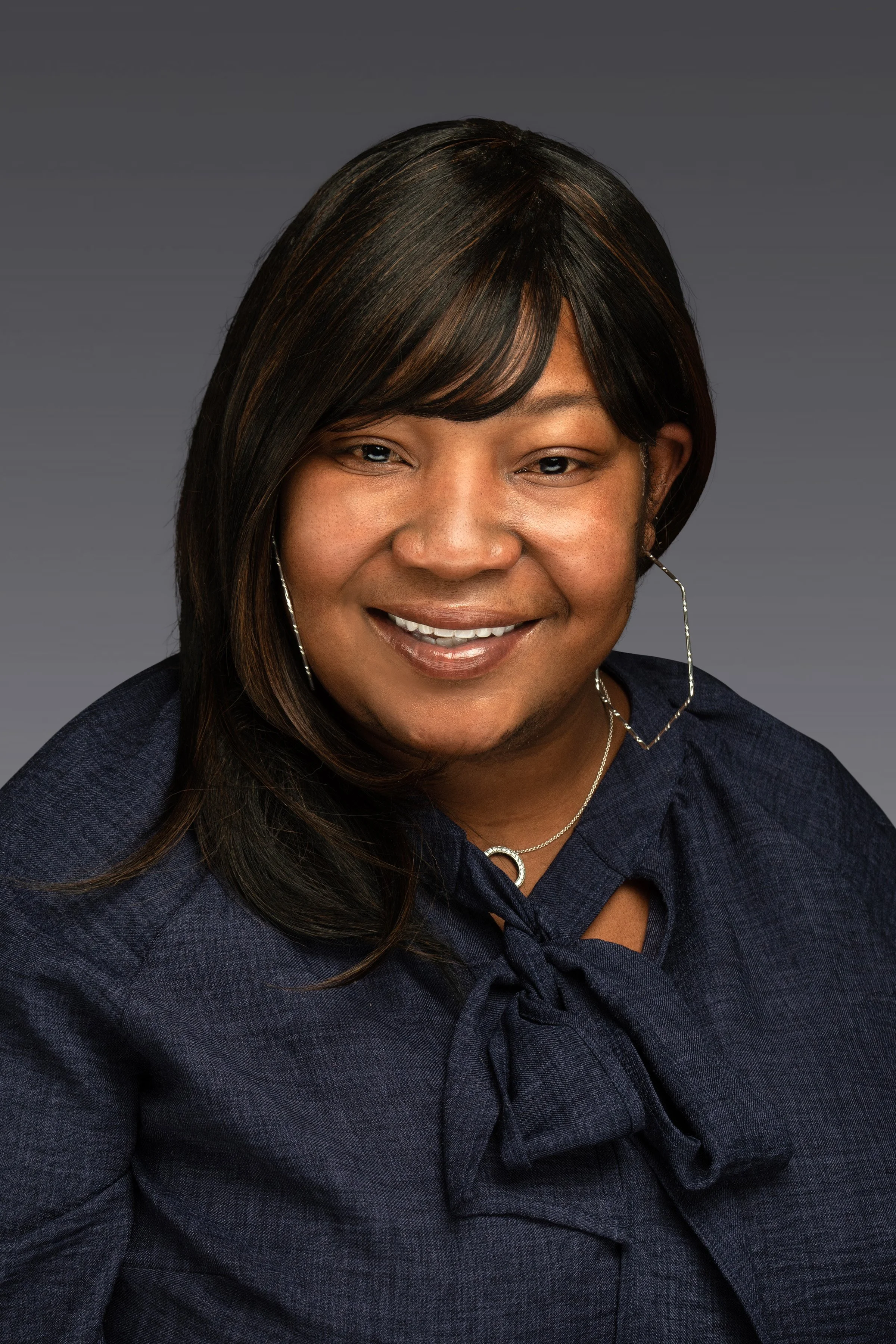 Smiling woman with dark hair wearing a navy blue blouse with a bow, silver hoop earrings, and a necklace against a gray background.