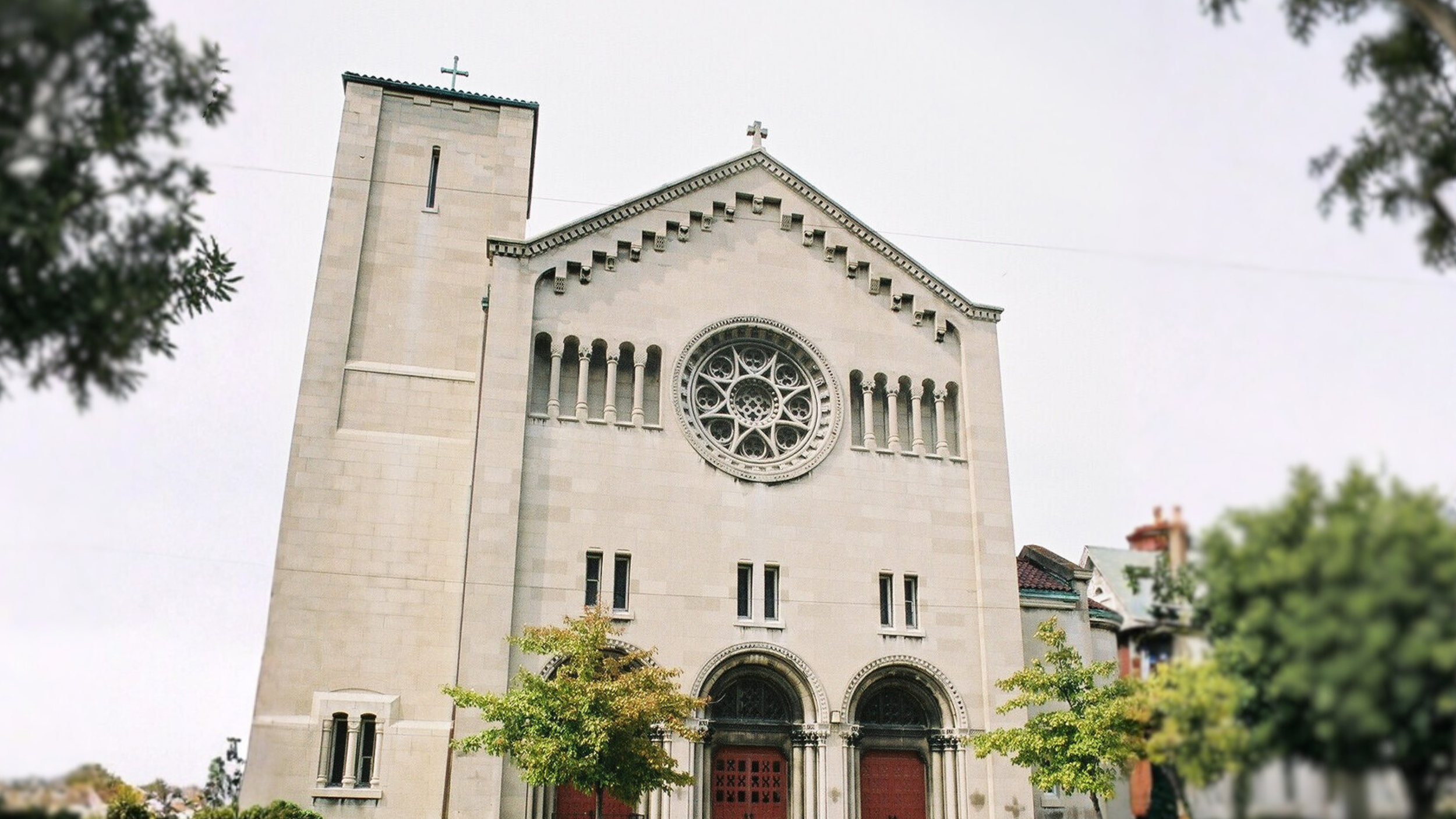 Photo of a church with a large rose window, stone facade, two arched doorways, and a tower with a cross on top, surrounded by green trees.