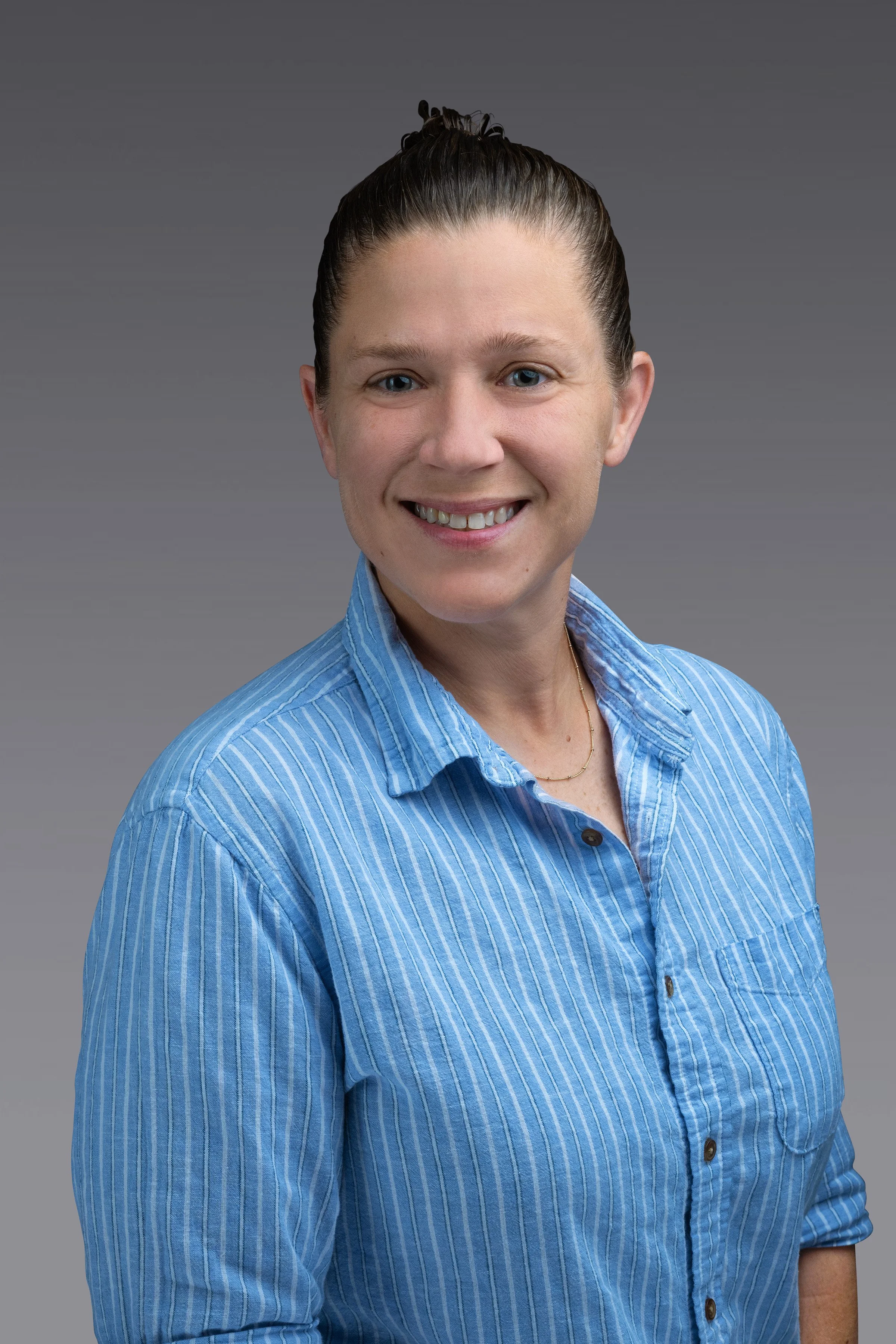 Portrait of a woman with dark hair in a bun, smiling, wearing a blue striped shirt, against a gray background.