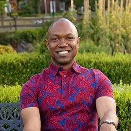 A smiling man sitting outdoors on a bench in a park, wearing a patterned red shirt with green trees and bushes in the background.
