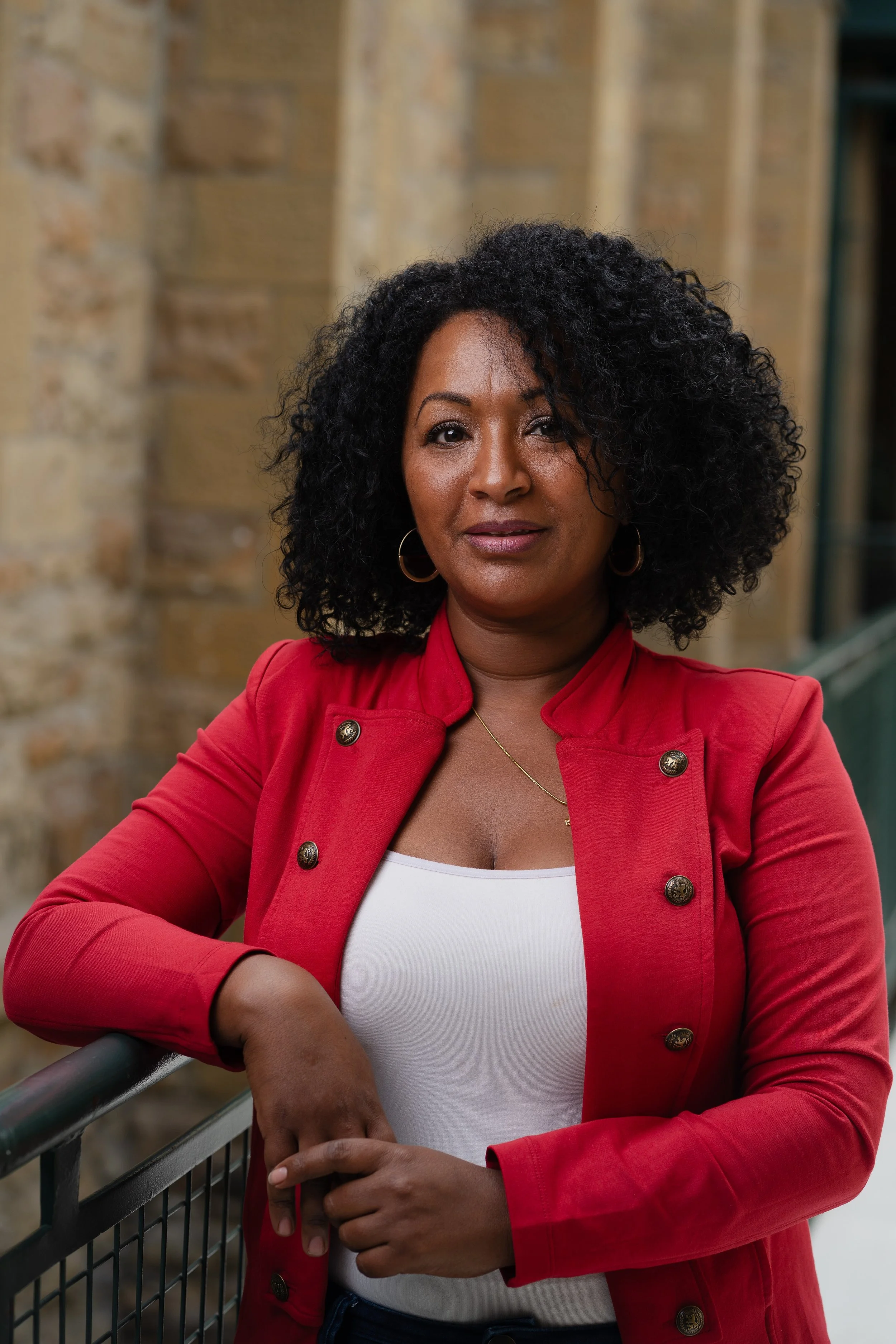 Woman with curly black hair wearing a red jacket and white top, standing outdoors with a stone wall background.