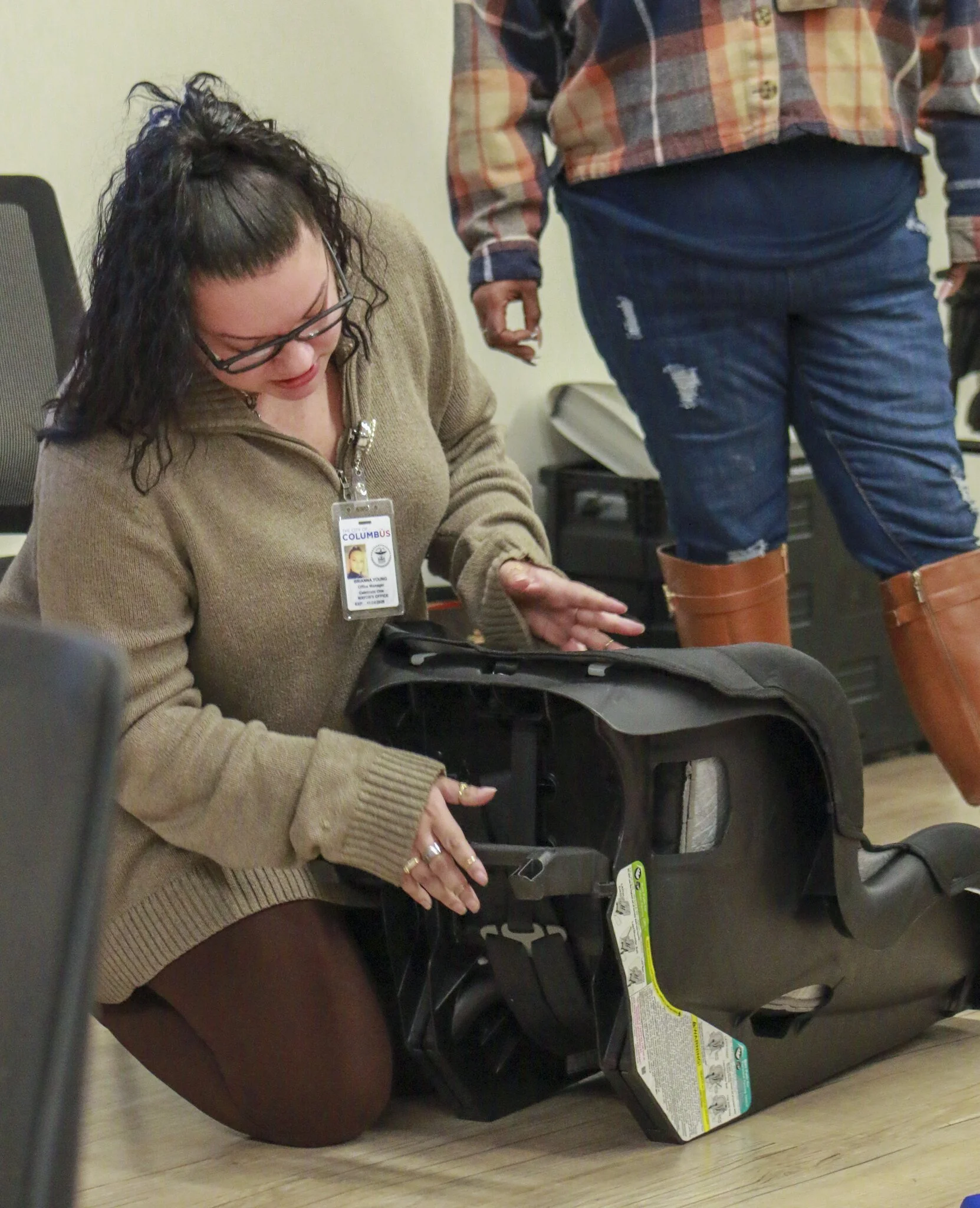 A woman in glasses kneeling on the floor, inspecting a car seat while a person in jeans and boots stands nearby.