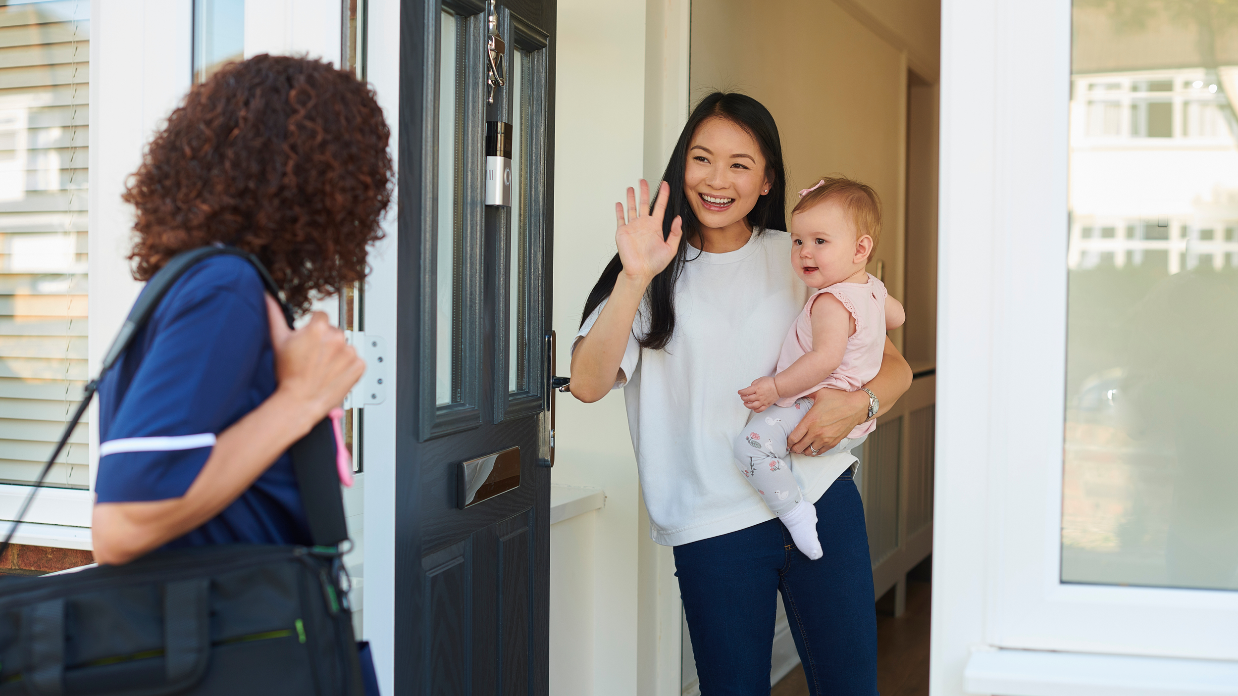 A woman holding a baby and waving hello to a delivery person at the front door of a house.