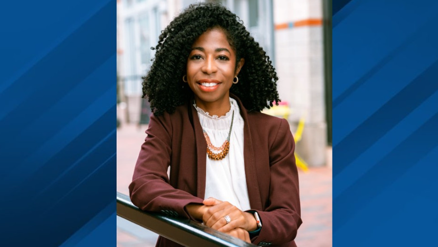 A woman with curly black hair, wearing a white blouse, brown blazer, and layered necklaces, sitting outdoors with a smile.