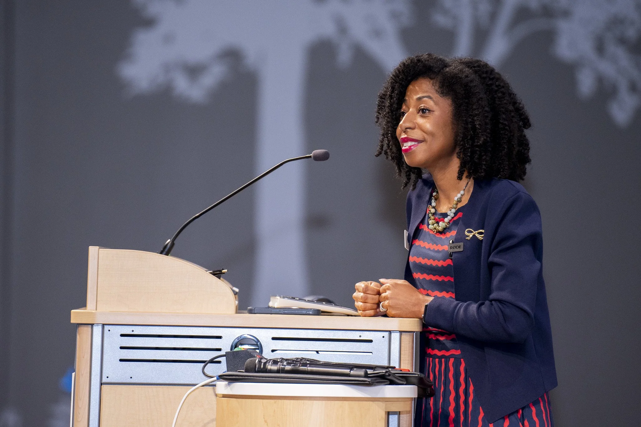 A woman speaking at a podium during a conference or event, with a background of a projection of a tree.