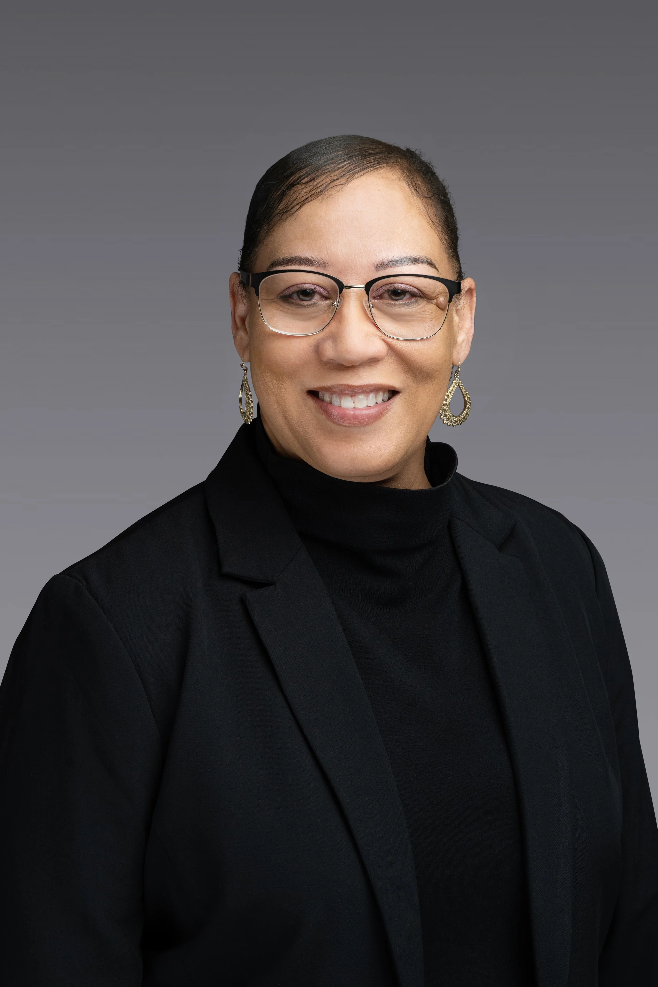A professional woman with glasses and earrings, smiling in front of a gray background.